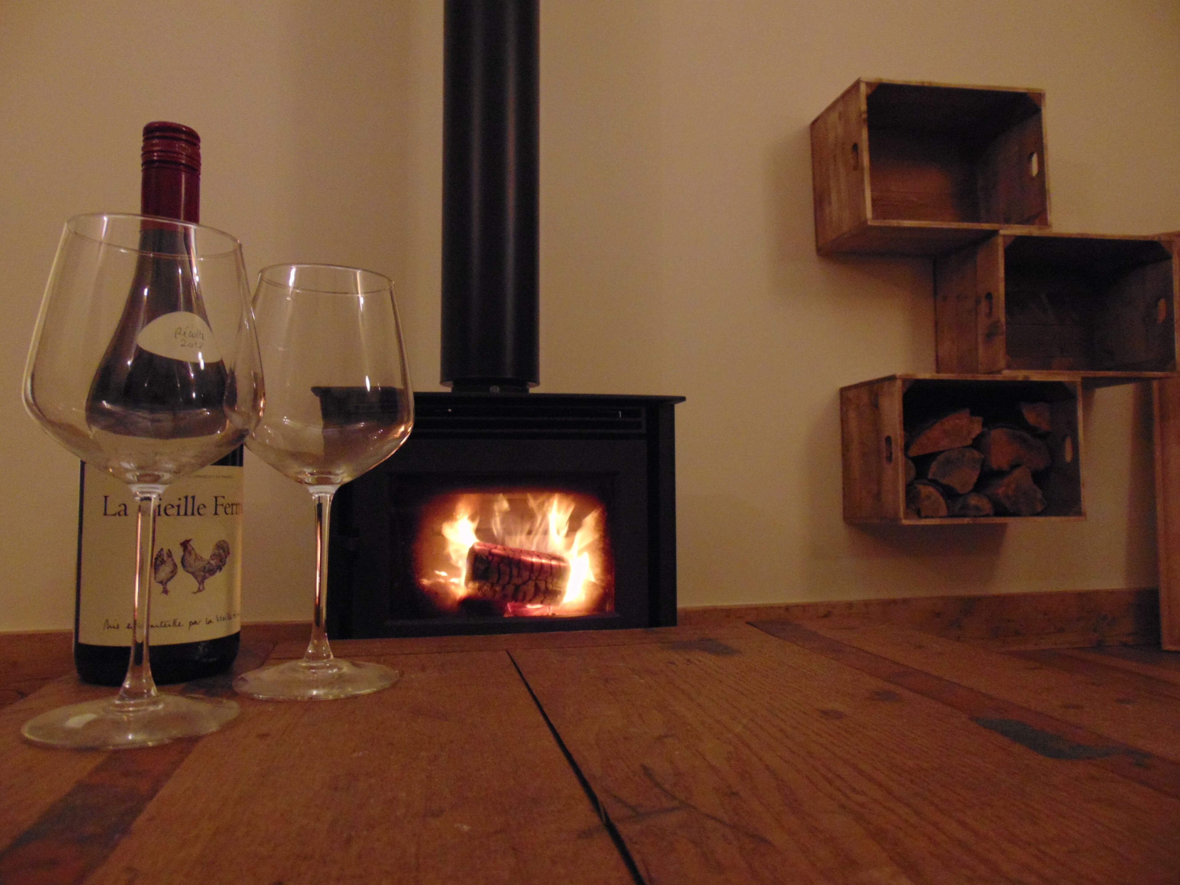 Una mesa de madera con dos copas de vino y una botella. Al fondo, una chimenea con leña encendida.
