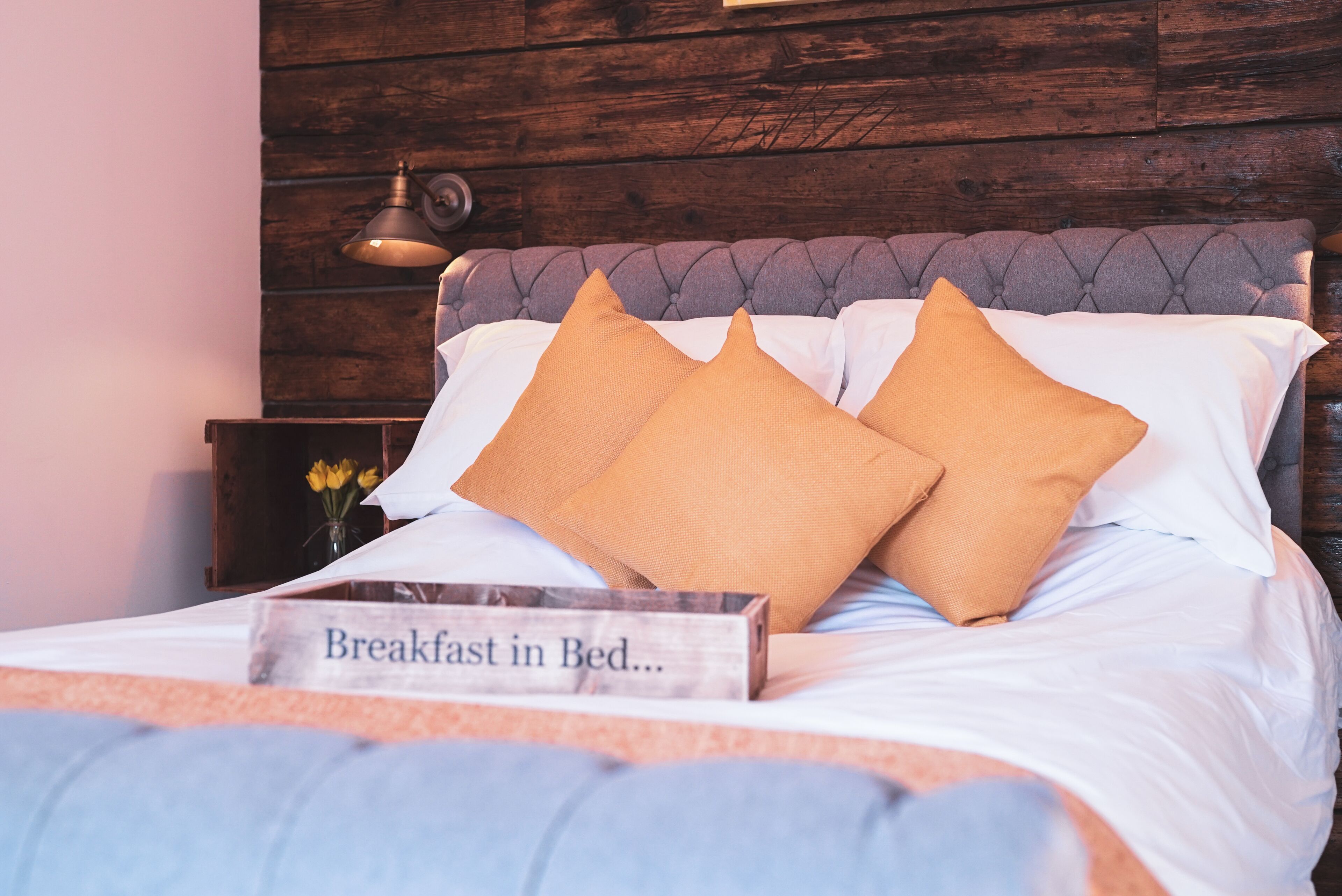 Cama bem arrumada com uma placa que diz 'Café da manhã na cama...' e cabeceira de madeira.