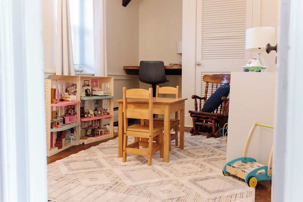 Une pièce avec une table et des chaises en bois, un petit bureau avec une chaise, une maison de poupée et une voiture jouet.