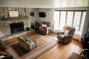 A living room with a stone fireplace, a sofa, a chair, a coffee table, and a large window with curtains.