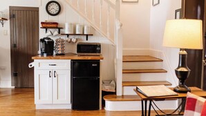 A kitchen with a microwave, coffee maker, and a small refrigerator. There is a wooden table with a lamp and a framed picture on the wall. - The Stone House Between the Streams (Gap)