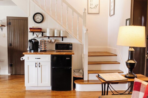 A kitchen with a microwave, coffee maker, and a small refrigerator. There is a wooden table with a lamp and a framed picture on the wall. - The Stone House Between the Streams (Gap)