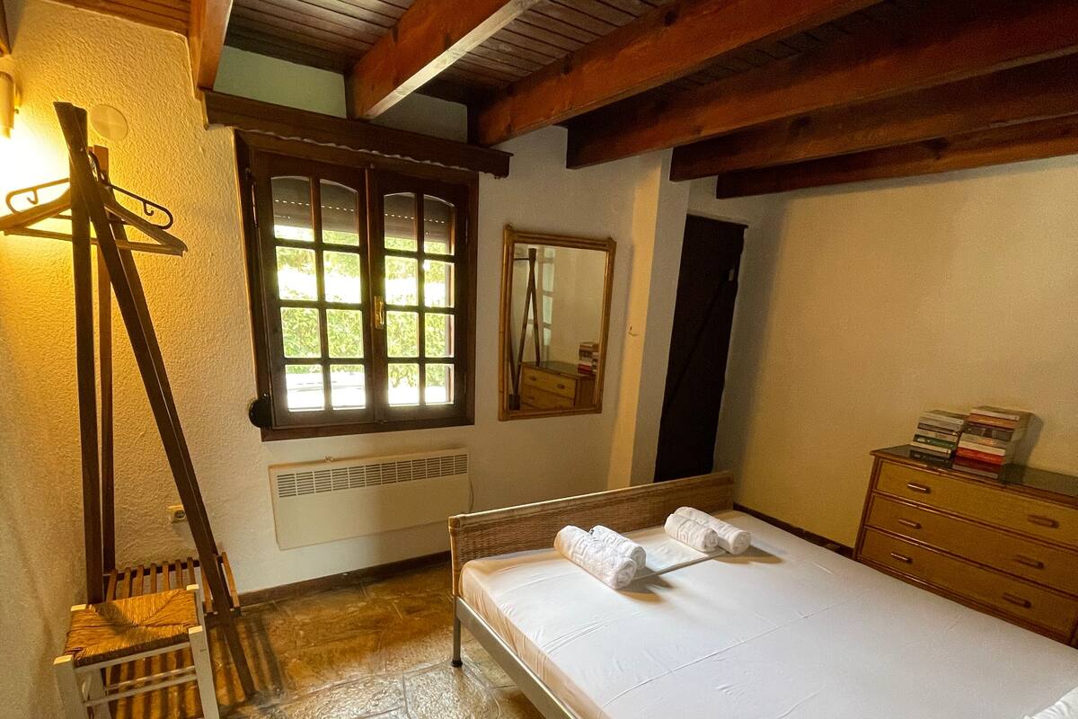 Ein Schlafzimmer mit einem Bett, einem Holzstuhl, einer Kommode, einem Spiegel und einem Fenster mit Blick auf Grünflächen.