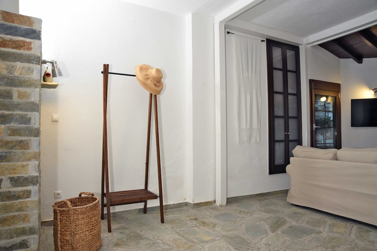 Ein Zimmer mit Steinboden, einer Holzbank, einem Hut auf einem Ständer, einem gewebten Korb und einem beigen Sofa.