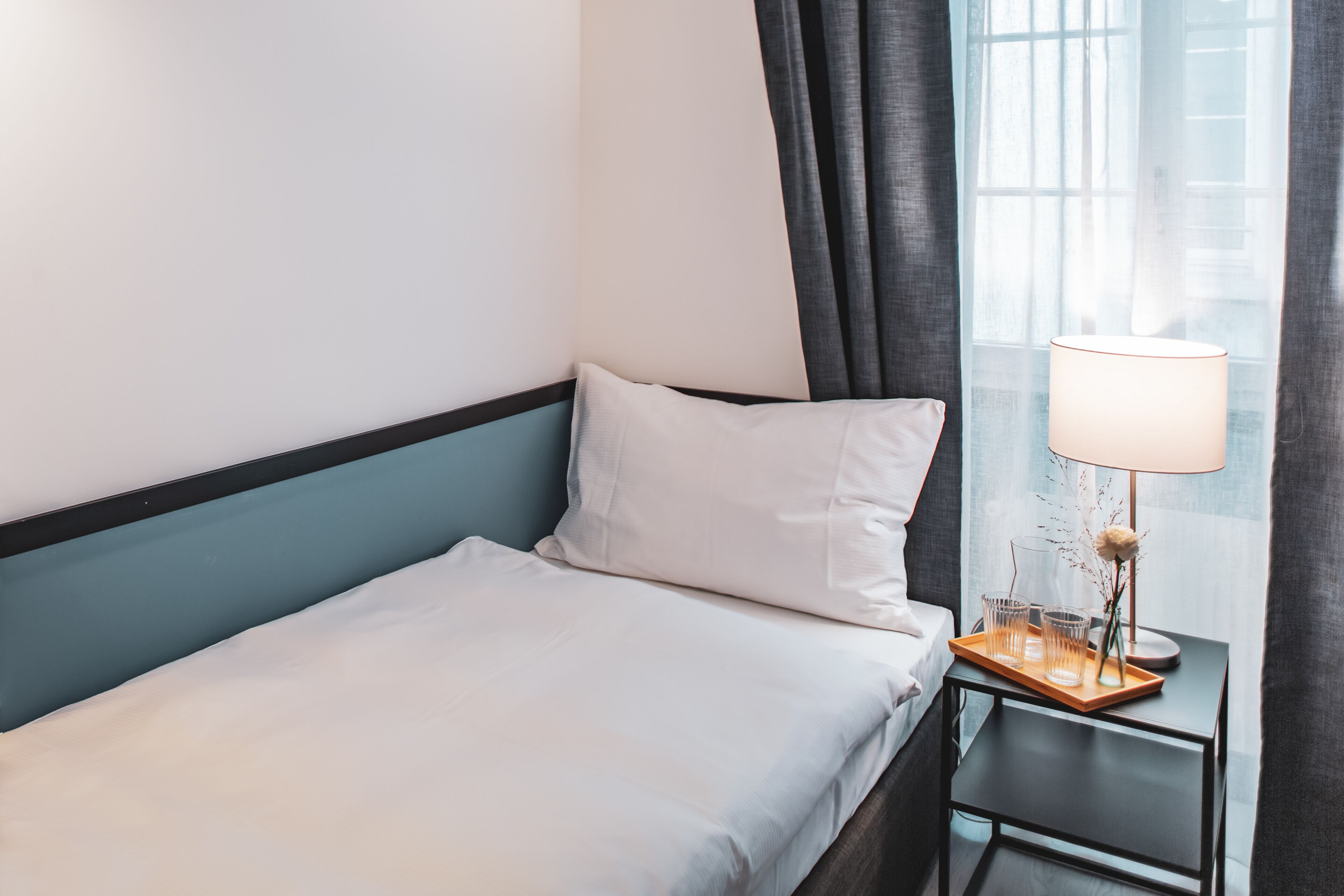 A neatly made bed with white linens, a bedside table with a lamp, and a window with curtains.