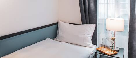 A neatly made bed with white linens, a bedside table with a lamp, and a window with curtains.