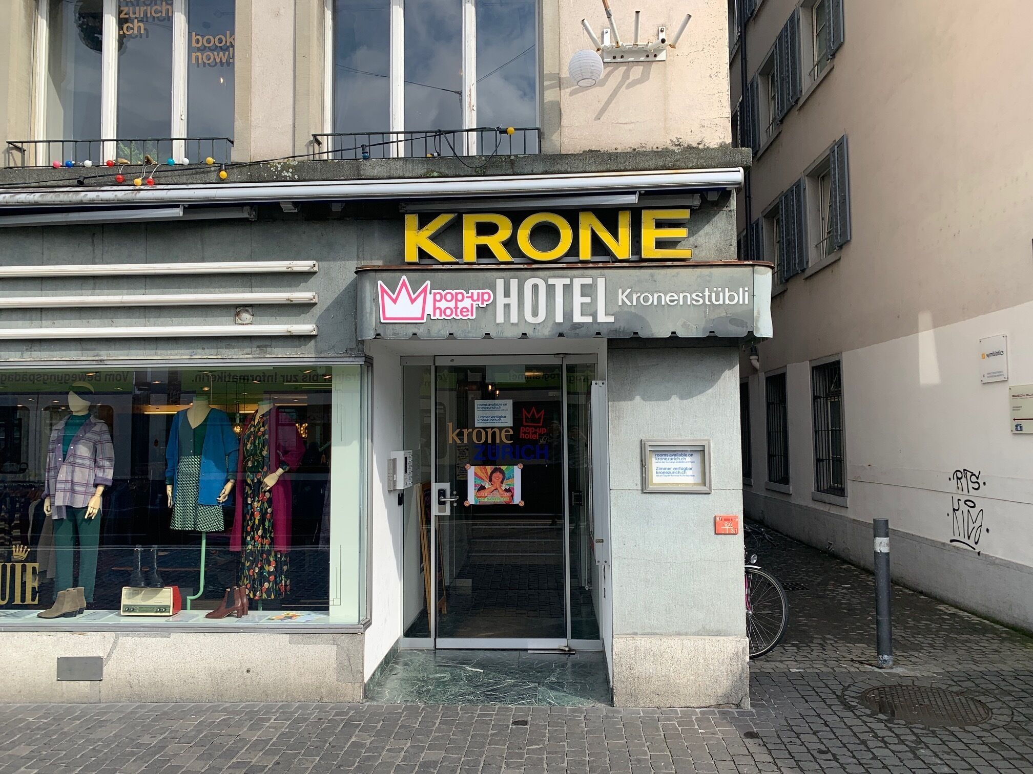Un hotel llamado "KRONE pop-up hotel" con una entrada visible y maniquíes mostrando ropa en el escaparate.