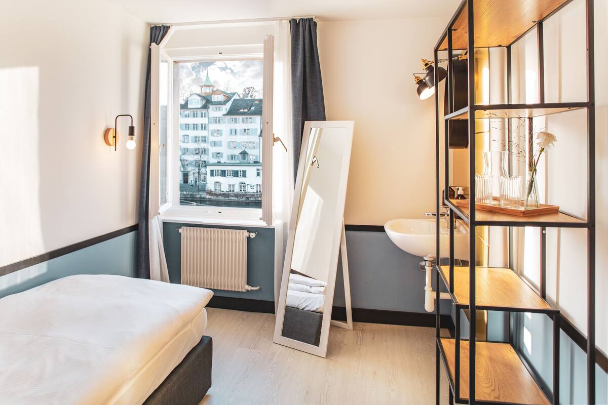 A hotel room with a bed, a window with a view of buildings, a mirror leaning against the wall, a sink, and a shelf with decorative items.