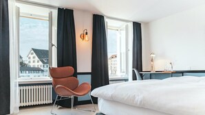 A modern hotel room with a large bed, a chair, a desk, and a view of buildings through large windows. - Pop Up Hotel Krone Zürich (Zürich)