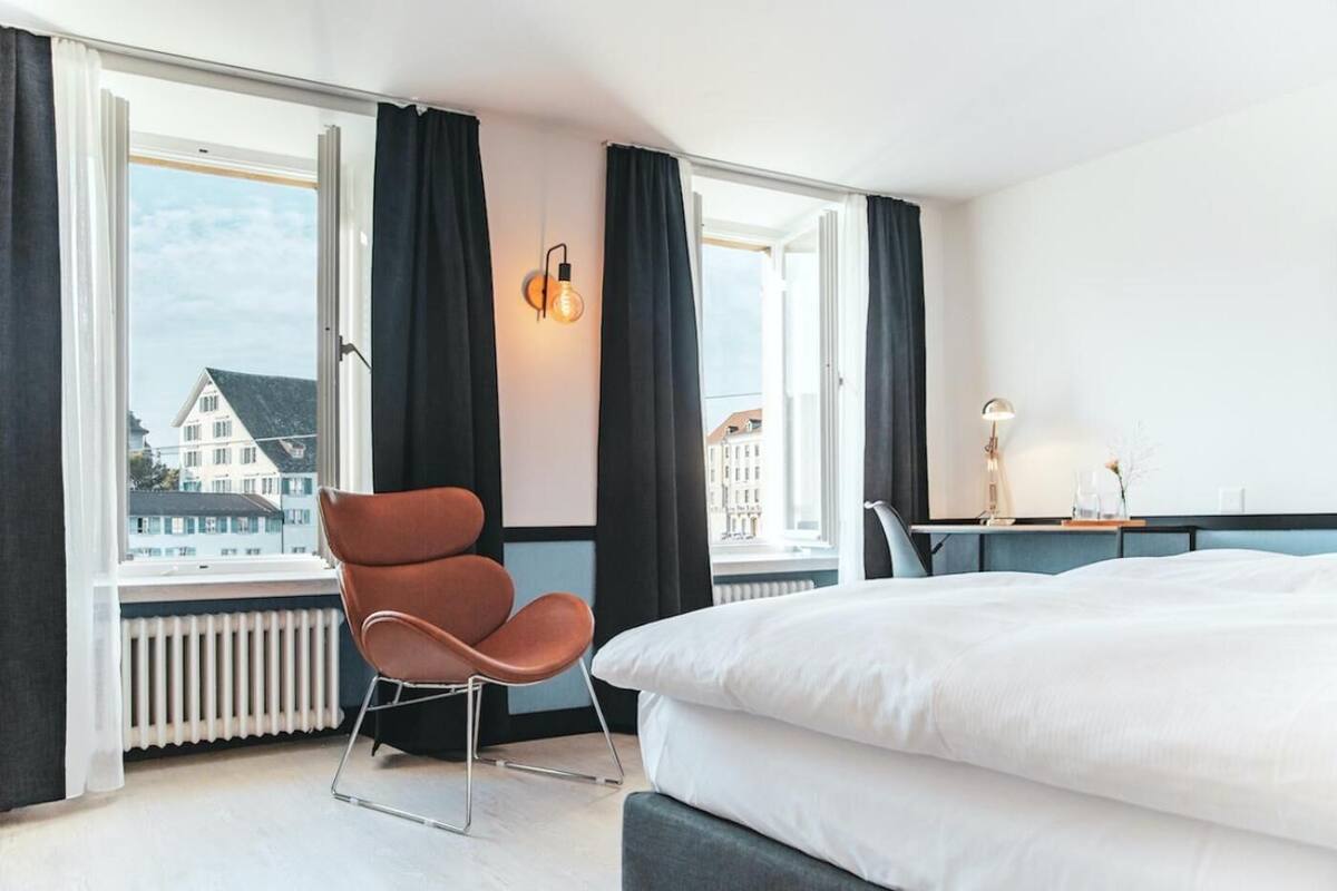 A modern hotel room with a large bed, a chair, a desk, and a view of buildings through large windows.