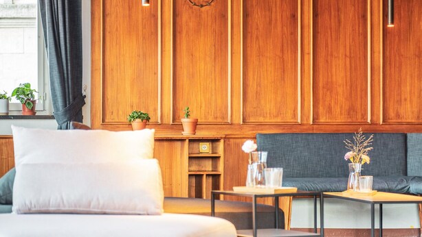 A hotel room with a wooden paneled wall, a bed with white bedding, a grey sofa, a small table with a vase of flowers, and a window with curtains. - Pop Up Hotel Krone Zürich (Zürich)