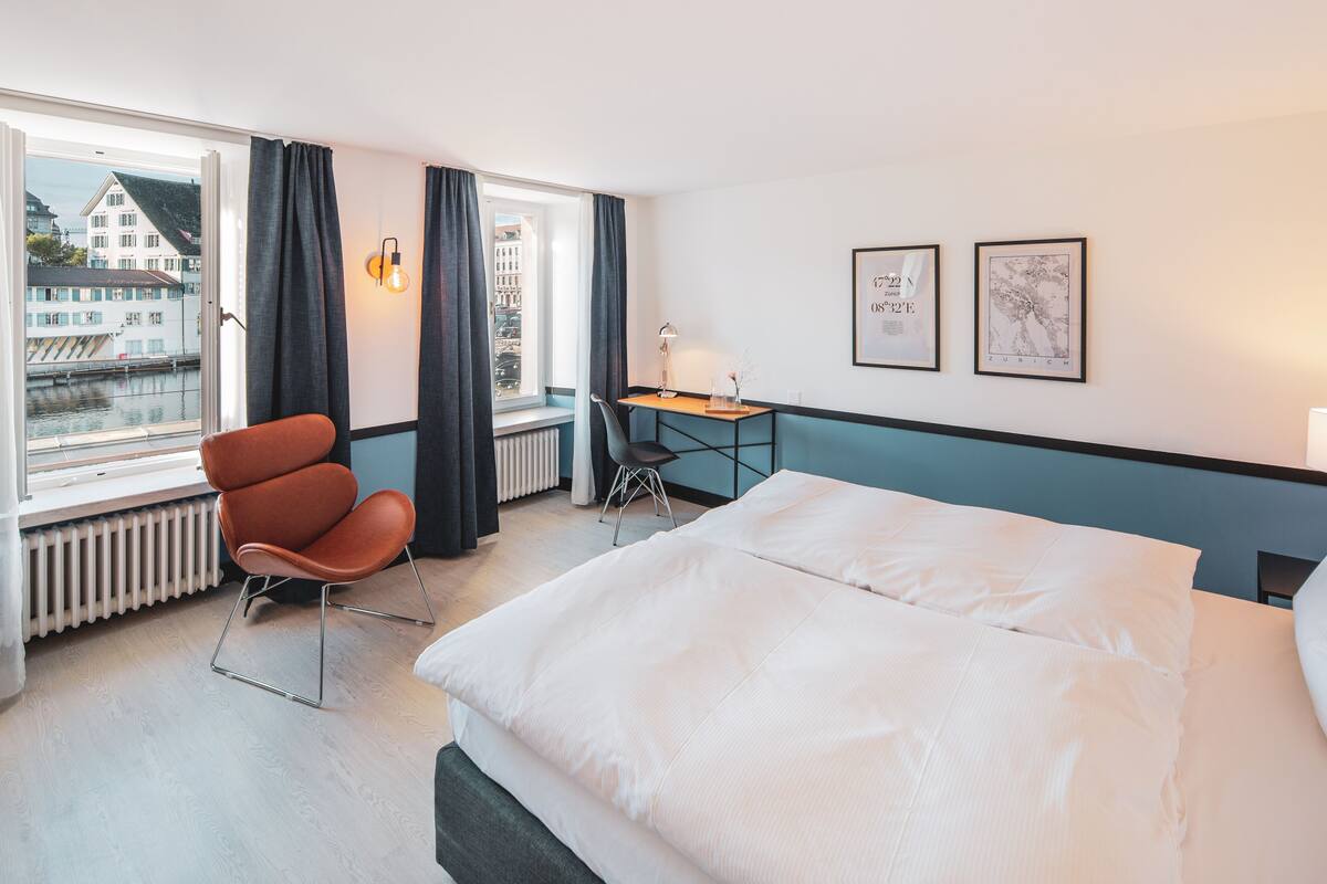 A hotel room with a bed, a desk, a chair, a window with curtains, and two framed pictures on the wall.
