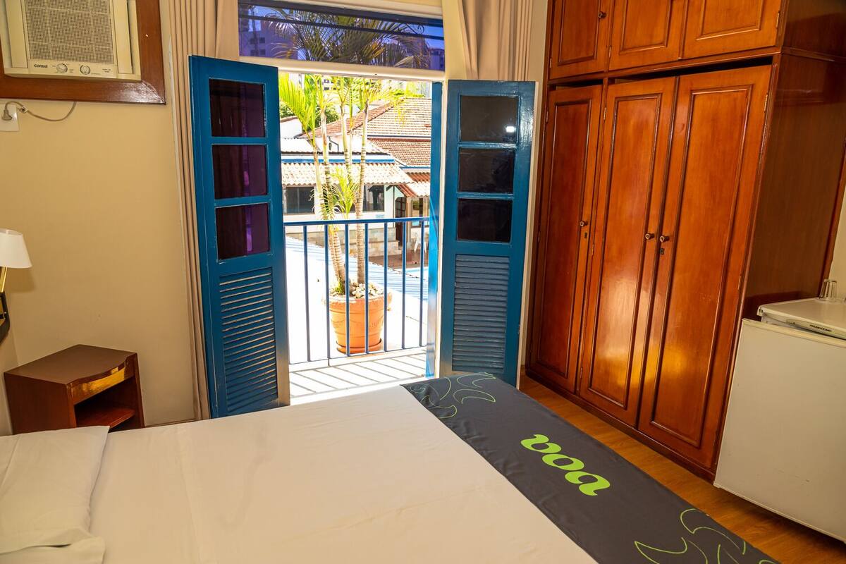 A hotel room with a bed, a desk, a wardrobe, and a balcony with a view of a building and palm trees.