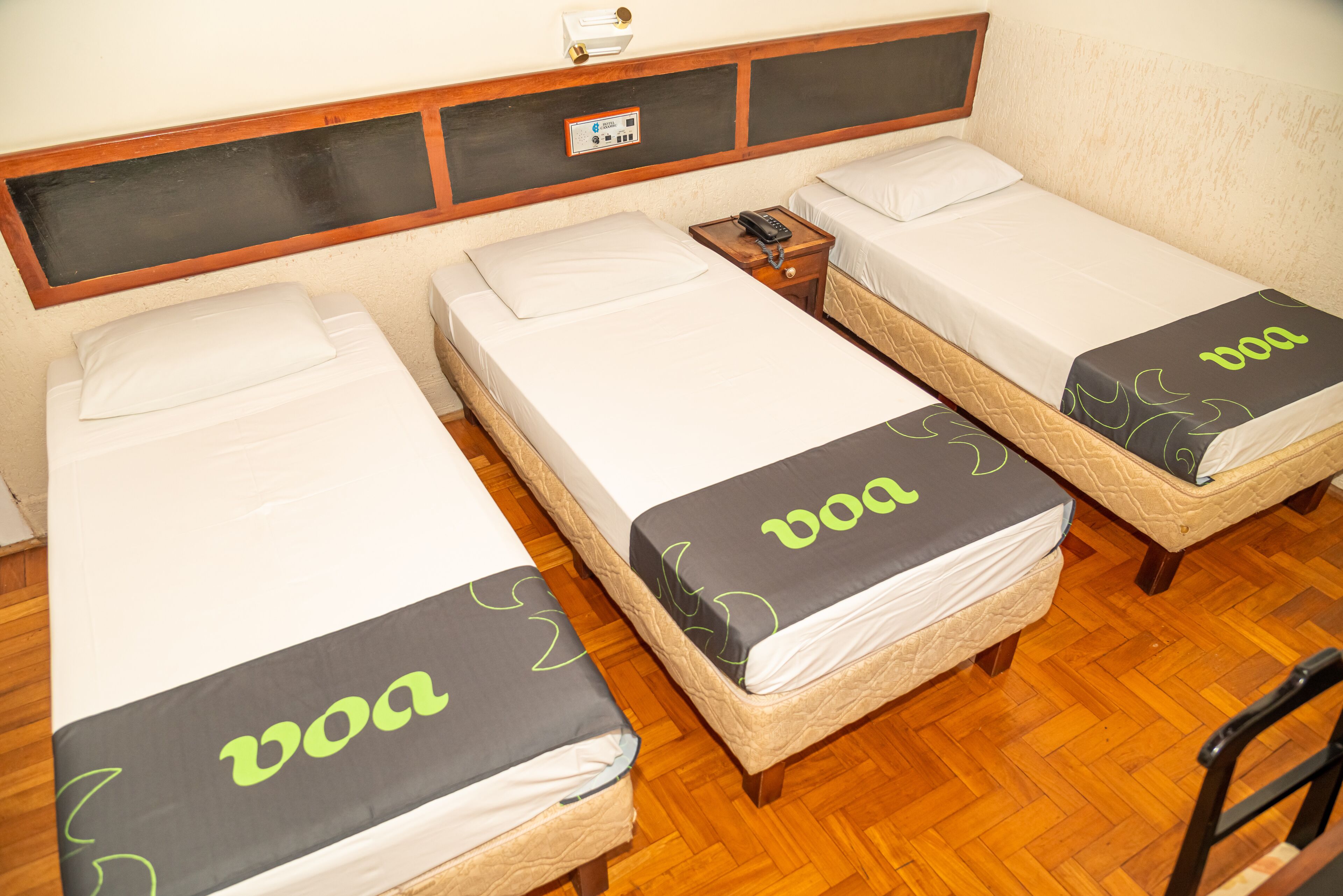 A room with three single beds, each with a 'VOA' branded bedspread. There is a bedside table with a phone on it, and a wall-mounted control panel above the beds.