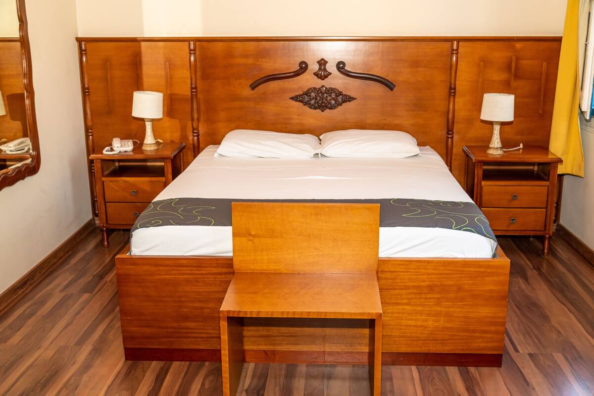 A wooden four-poster bed with a matching nightstand on each side, a bedside table with a lamp, and a wooden chair in front of the bed.