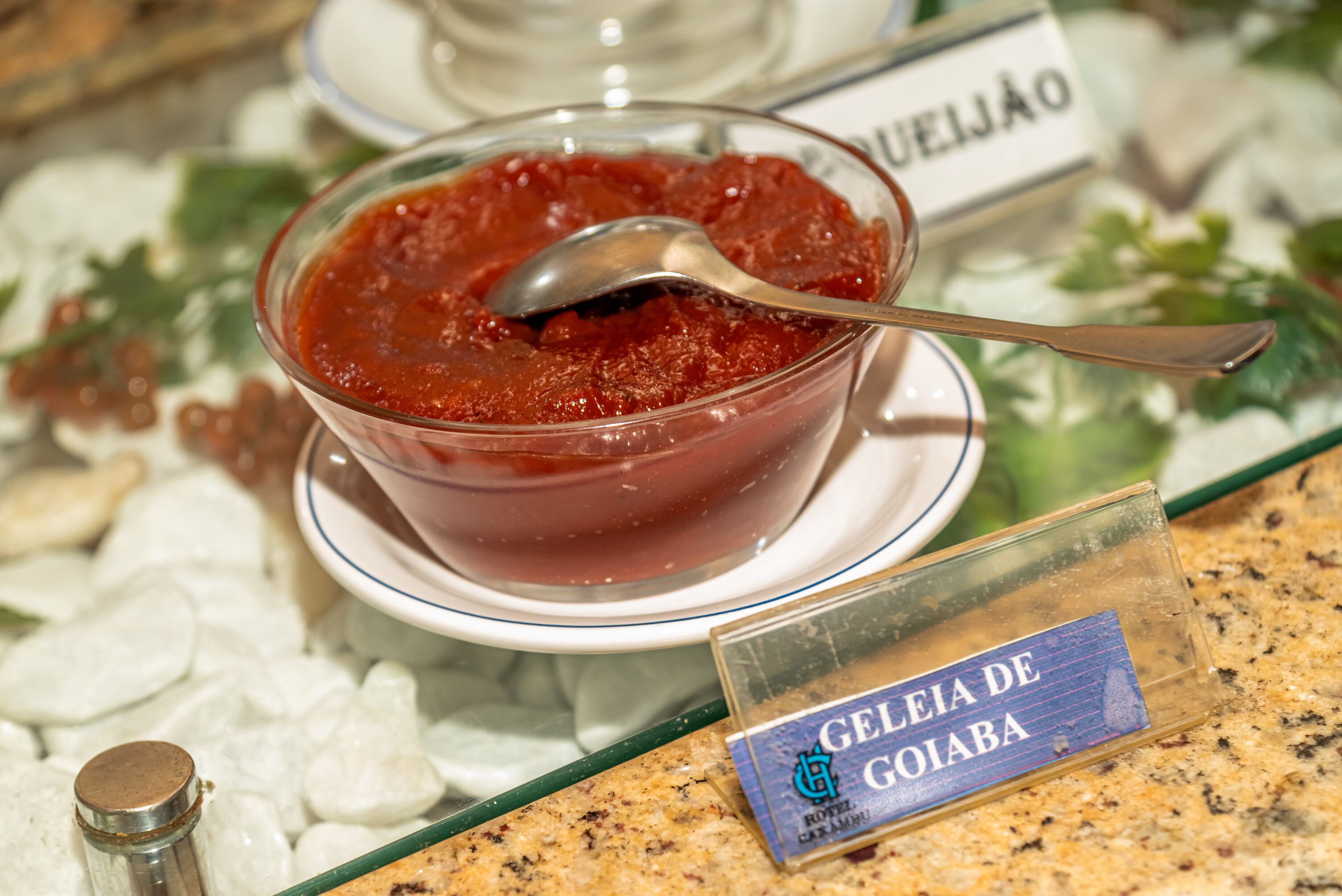A bowl of red jelly with a spoon, labeled "Geleia de Goiaba," on a table with a name tag.