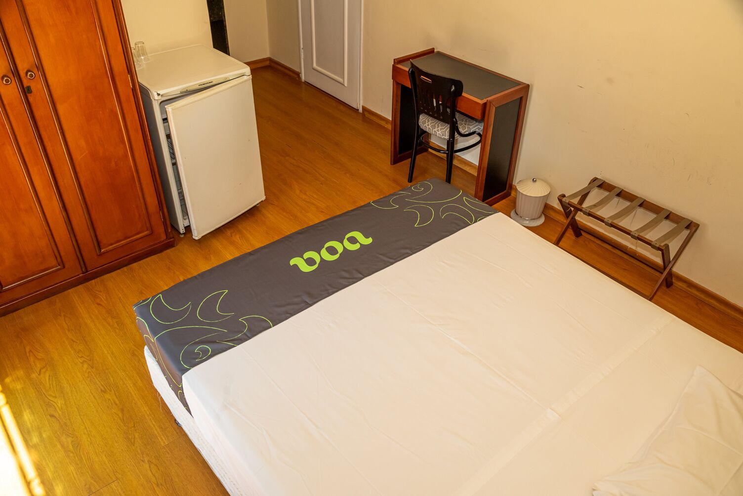 A hotel room with a bed, a small desk, a chair, a refrigerator, and a waste bin.