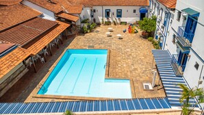 A swimming pool with a tiled deck, surrounded by buildings with red-tiled roofs and blue accents. - VOA Hotel Caxambu (Caxambu)