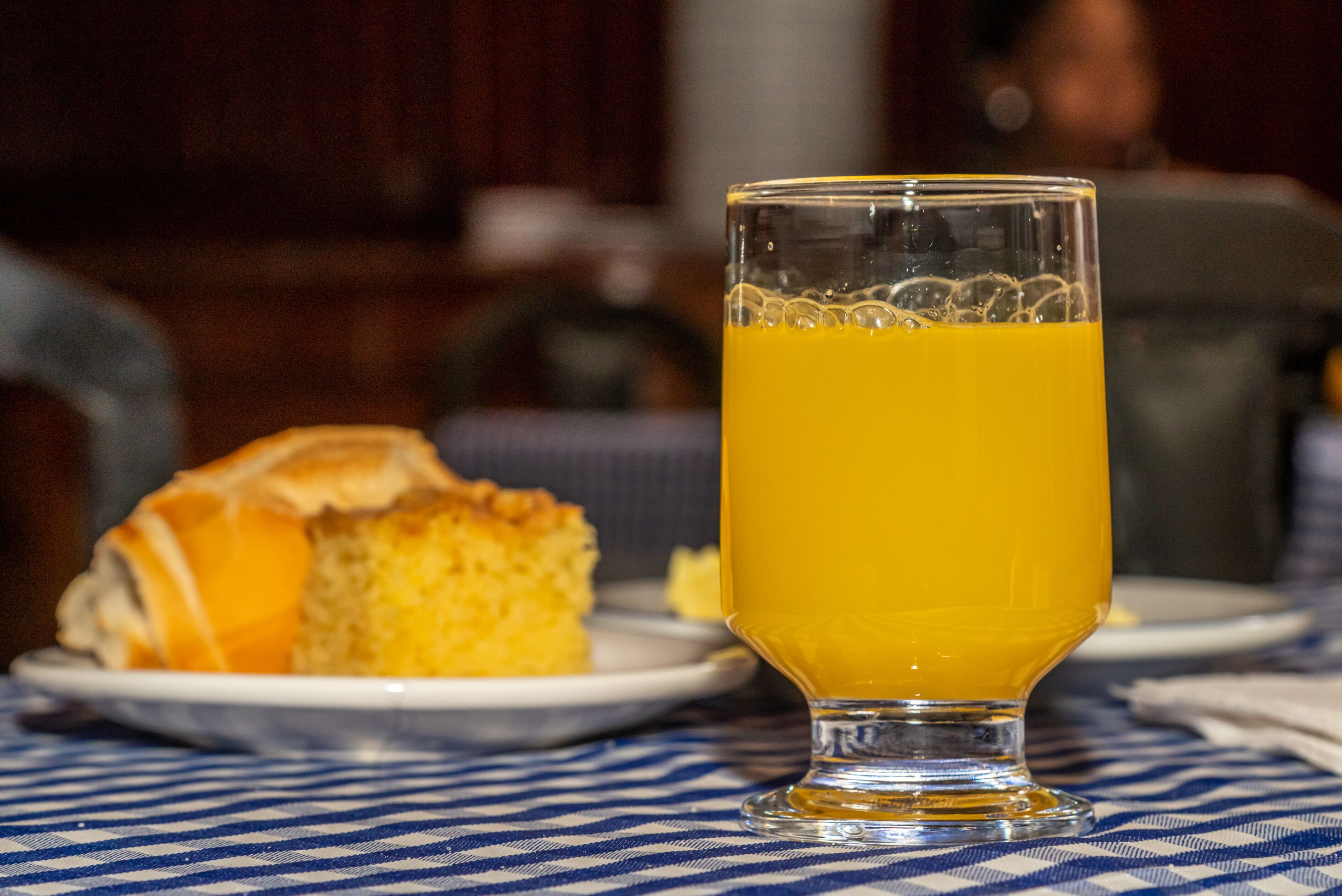 A glass of orange juice on a checkered tablecloth with a slice of cake on a plate.