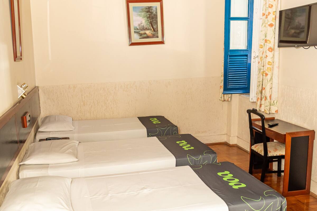 A hotel room with three single beds, a wooden ceiling, a desk with a chair, a television, and a window with blue shutters.