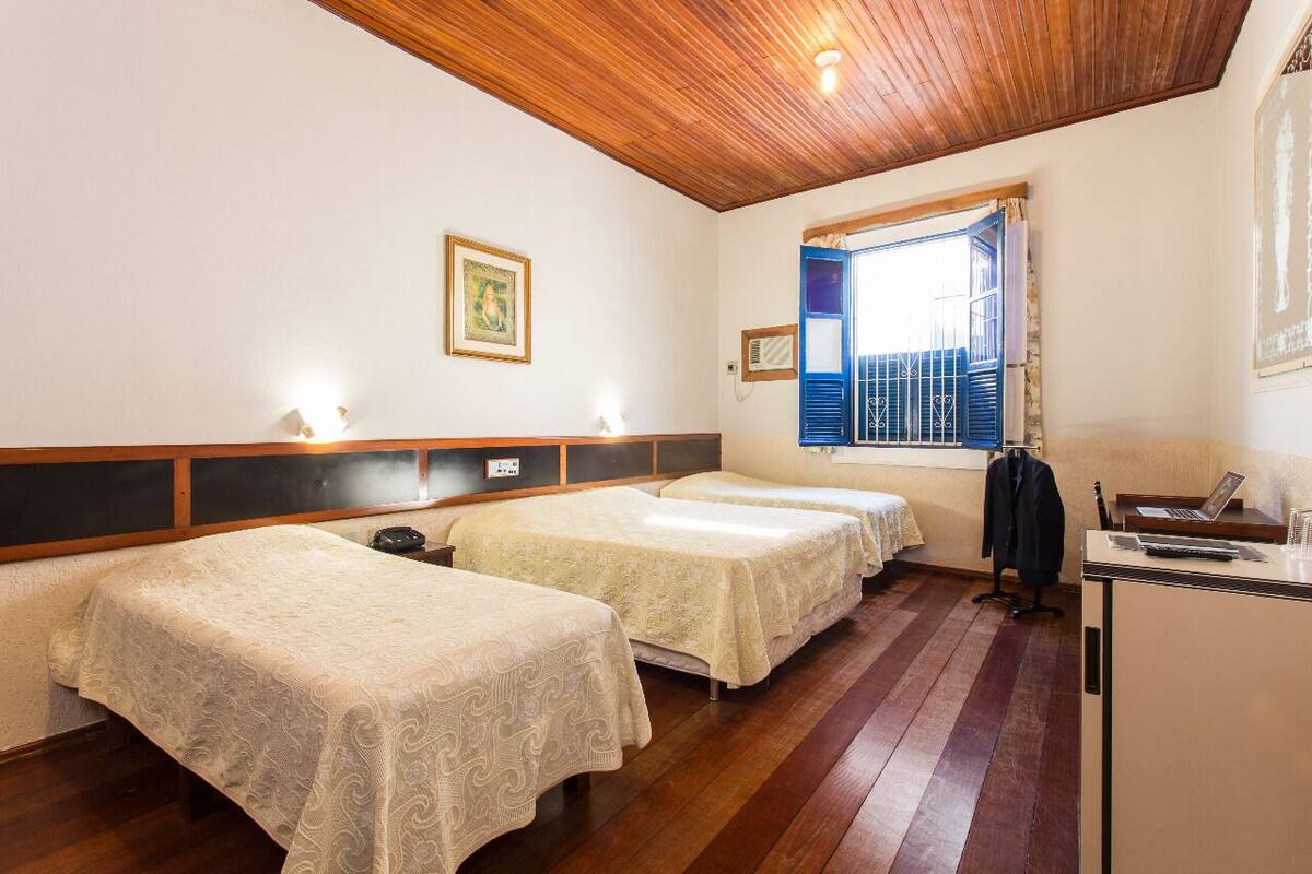 A room with two single beds, a wooden ceiling, a small desk with a lamp, and a window with blue shutters.