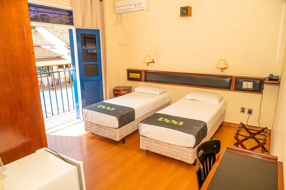 A hotel room with two single beds, a wooden desk, a chair, a small table, and a balcony with a railing.