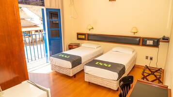 A hotel room with two single beds, a wooden desk, a chair, a small table, and a balcony with a railing.