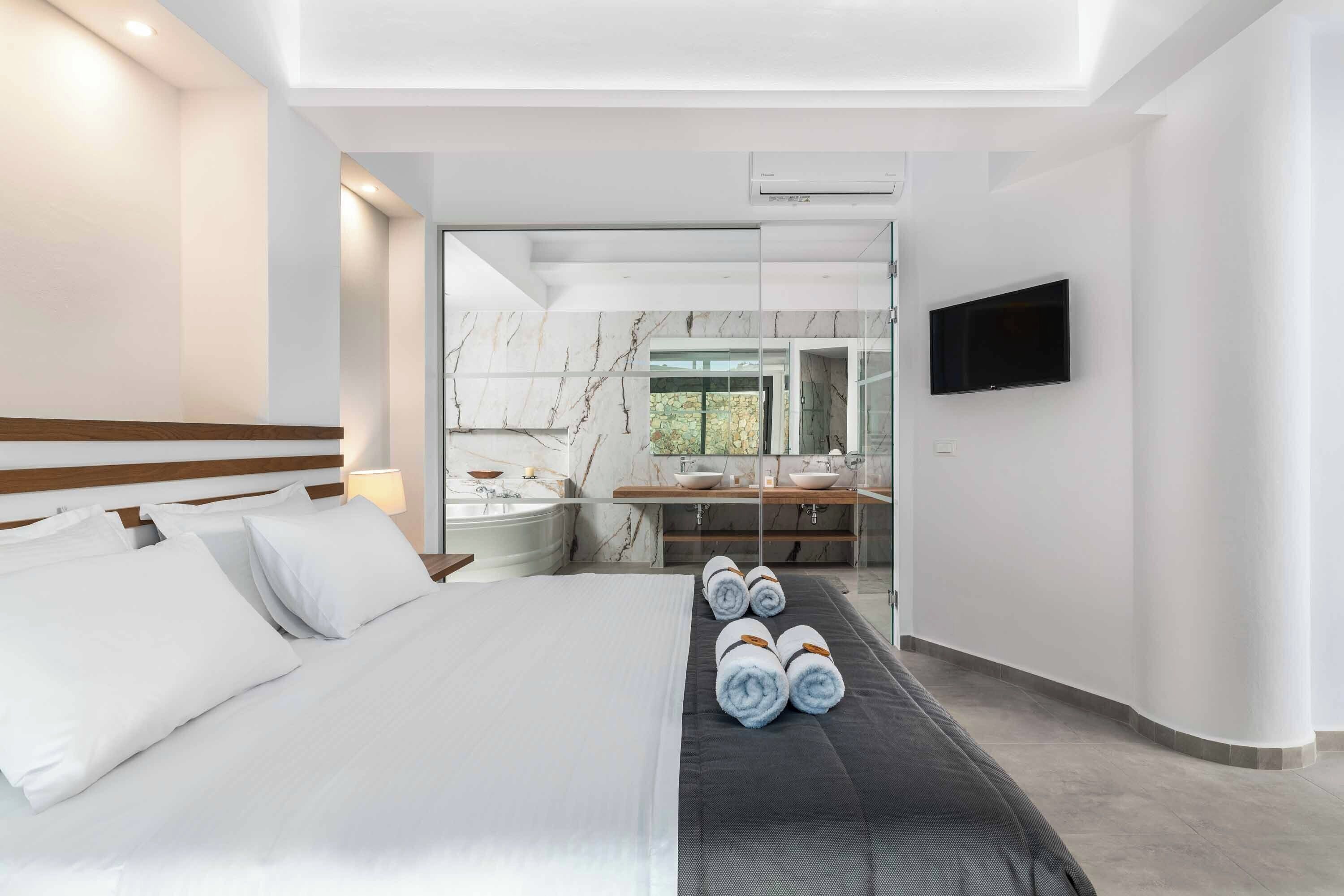 Moderni hotellihuone, jossa on suuri sänky, lasiseinäinen kylpyhuone, taulutelevisio ja näkymä ulos.