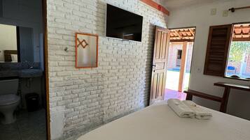 A room with a white brick wall, a bed with a white bedspread, a flat-screen TV mounted on the wall, and a wooden door leading to an outdoor area.