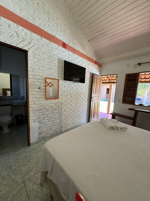 A room with a white brick wall, a bed with a white bedspread, a flat-screen TV mounted on the wall, and a wooden door leading to an outdoor area. - Pousada Askalon (Itaunas)