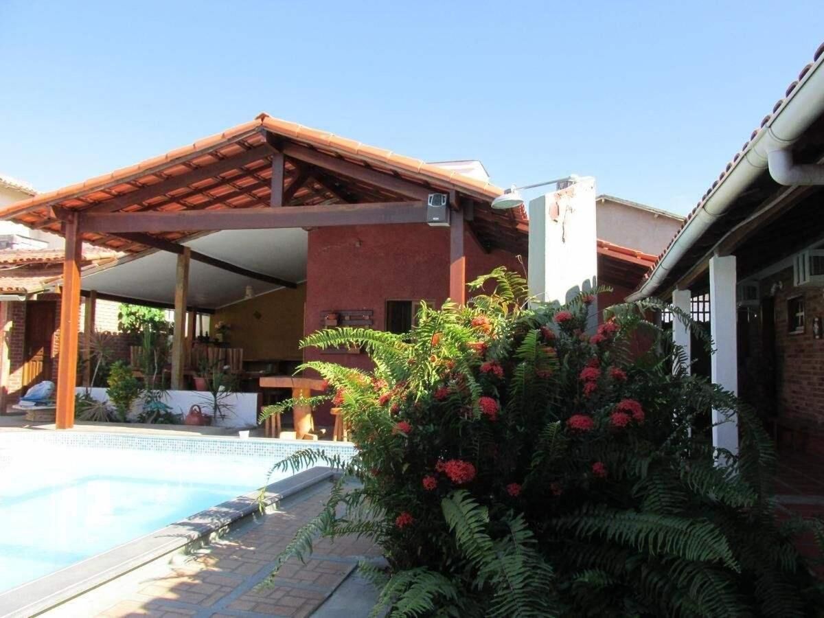 Un área junto a la piscina con un porche cubierto, un edificio de ladrillo y abundante vegetación.