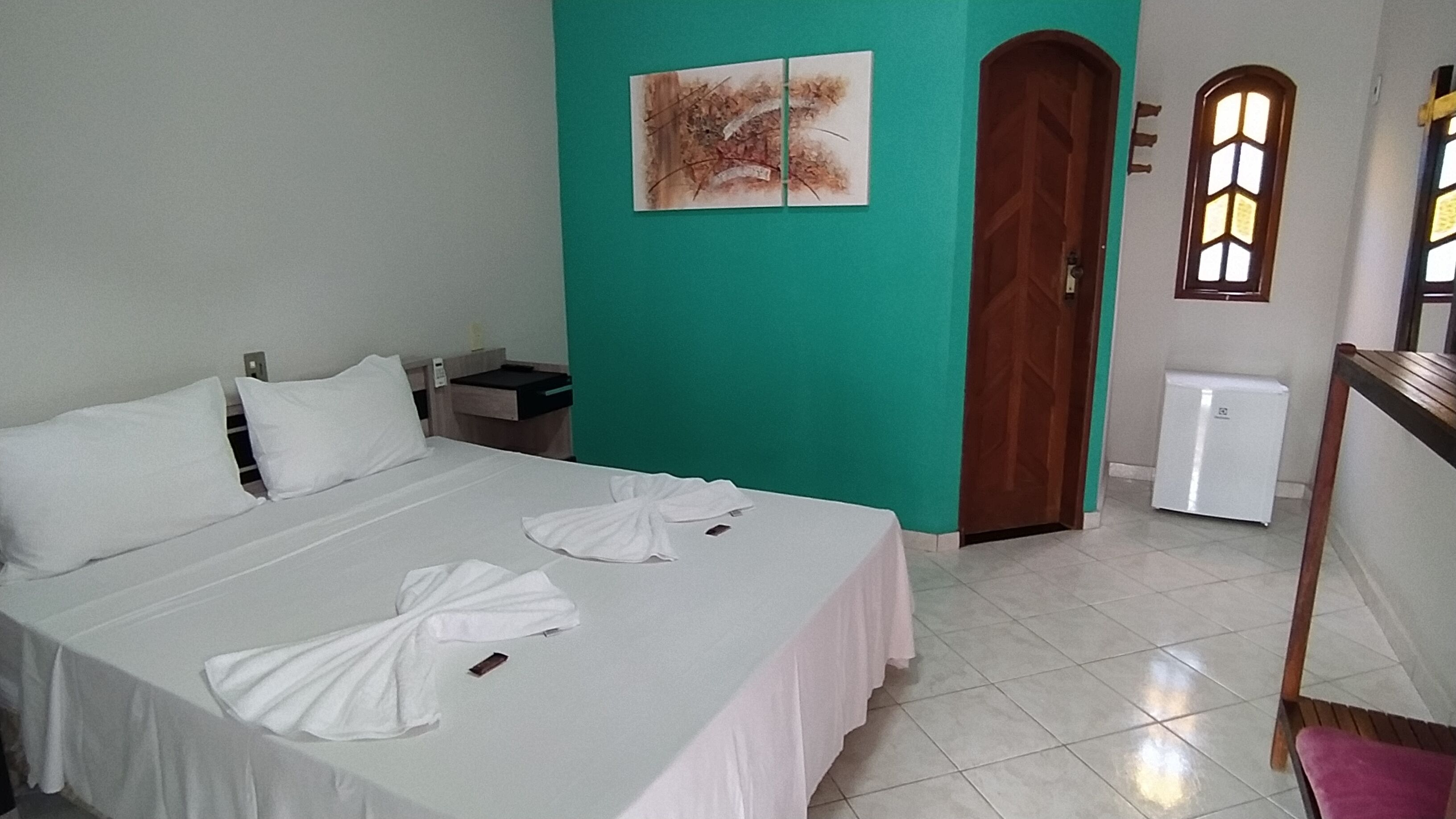 A hotel room with a bed, a nightstand, a wall-mounted painting, a door, and a window.