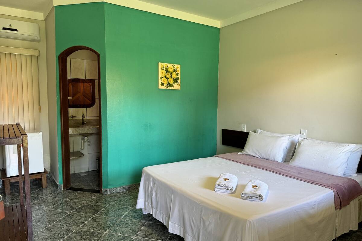 Ein Hotelzimmer mit Bett, kleinem Schreibtisch, Stuhl und einem Badezimmer mit Spiegel, sowie einer grünen Wand.