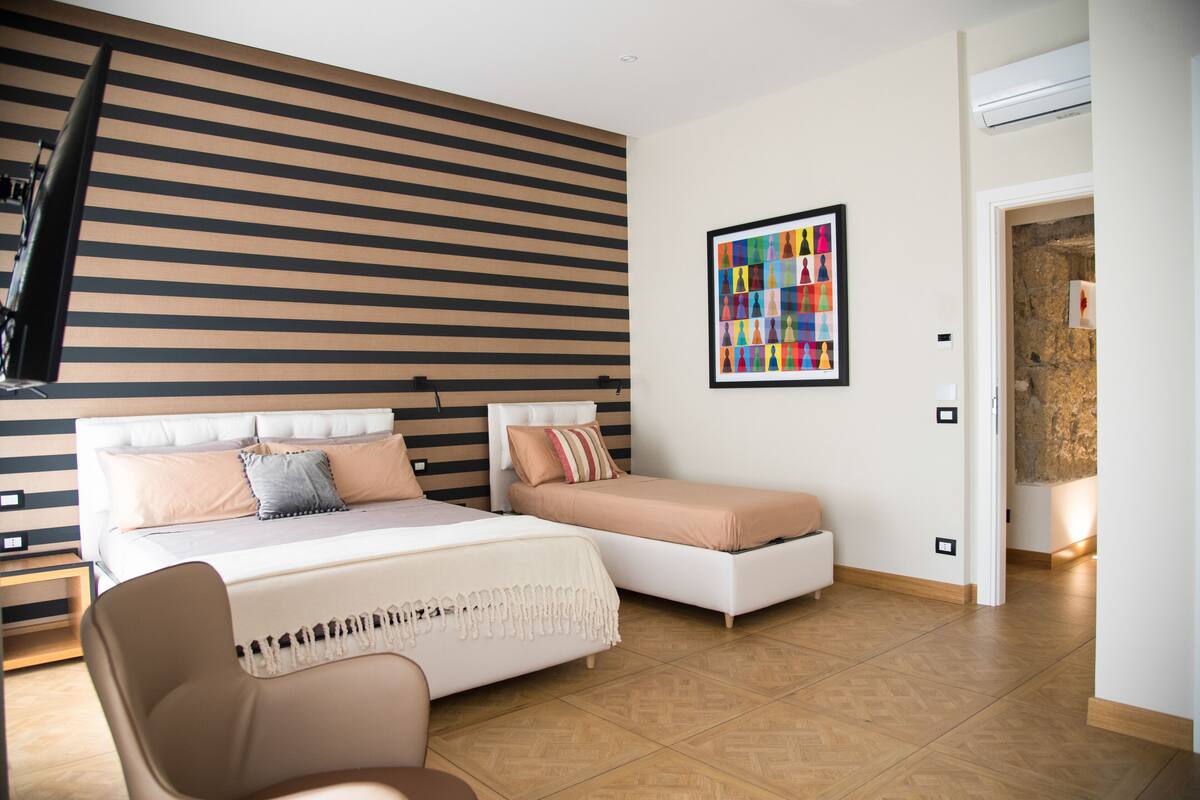 A modern bedroom with a large bed, a smaller bed, a chair, and a colorful artwork on the wall.