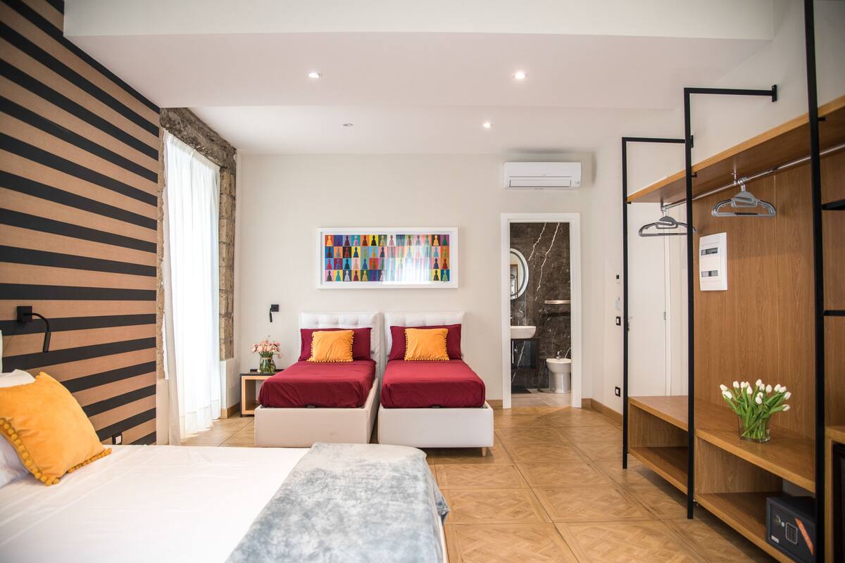 A modern bedroom with two beds, a wooden wardrobe, a bathroom with a marble wall, and a colorful artwork on the wall.