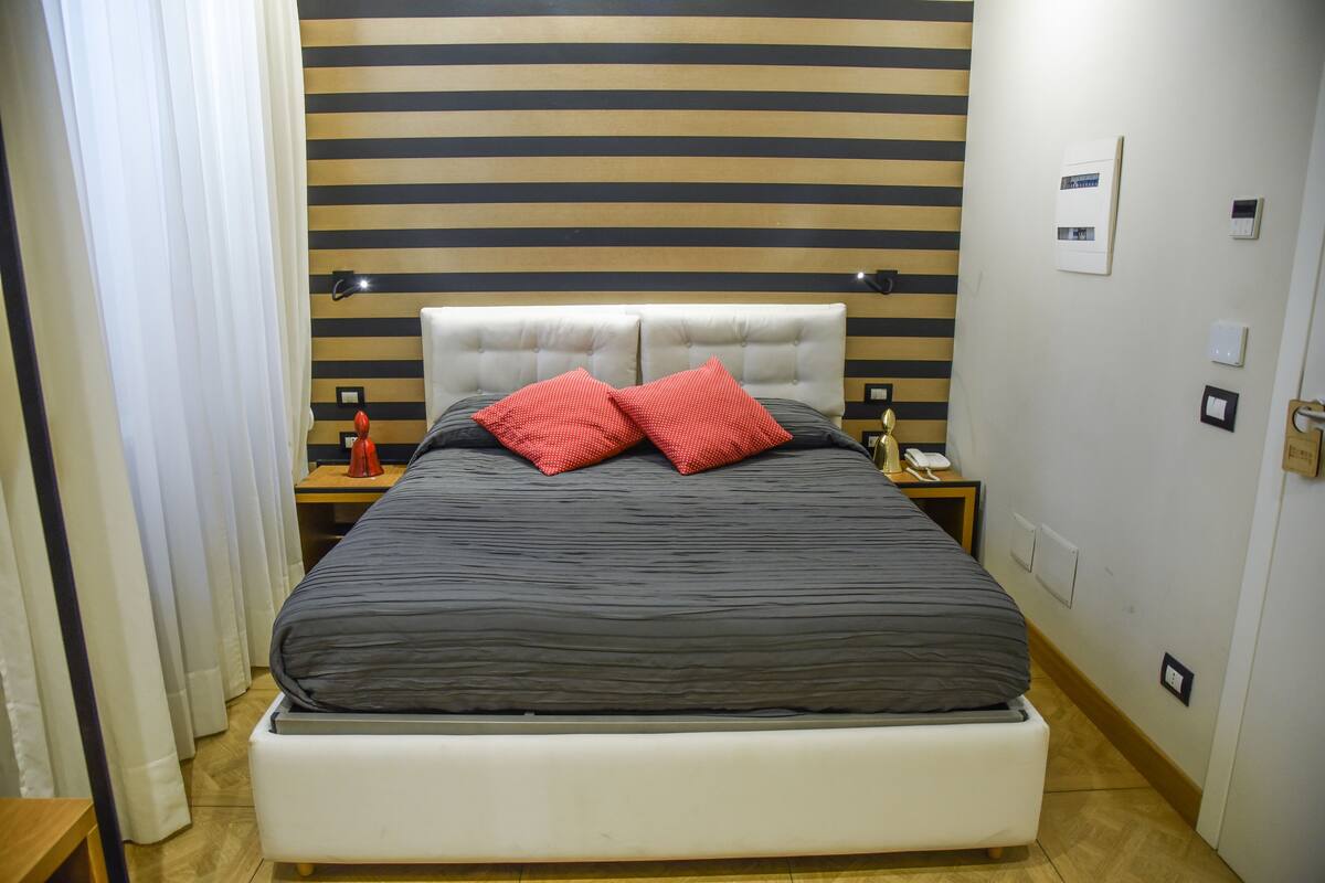 A neatly made bed with a grey and black striped headboard, two red pillows, and bedside tables with lamps and a telephone.