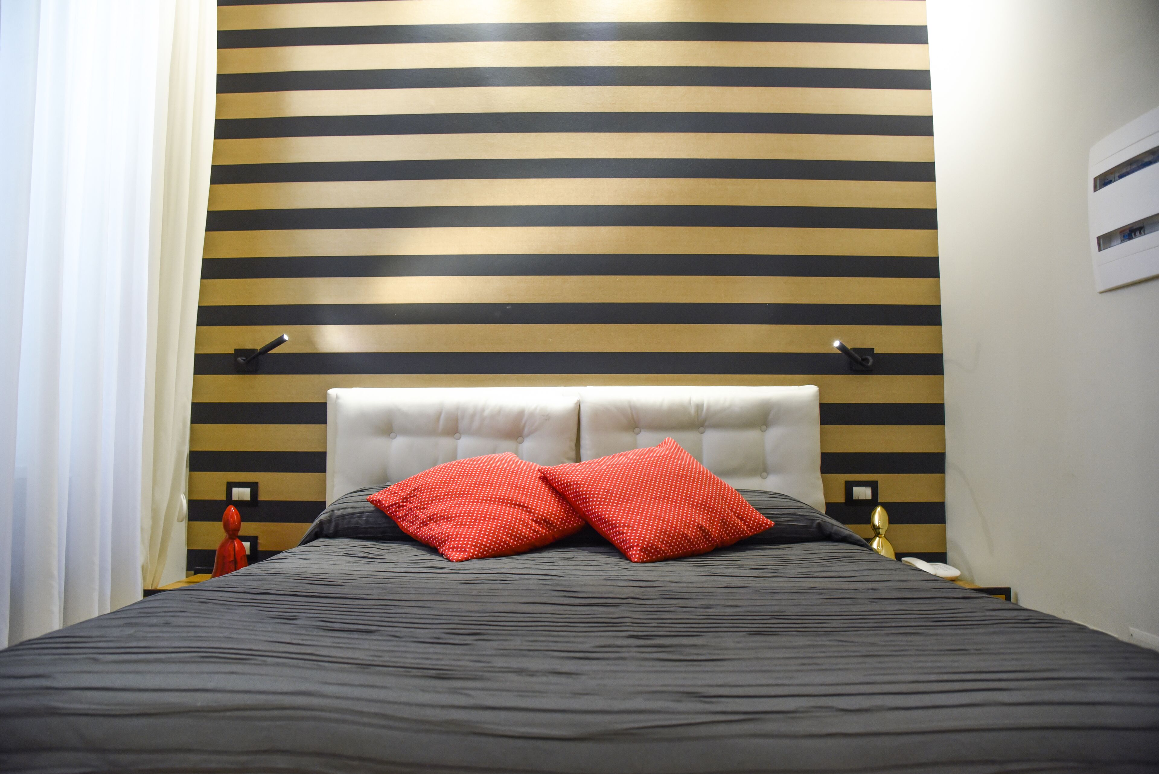 A hotel room with a striped wall, a tufted headboard, and two red pillows.