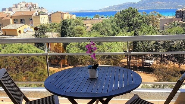 Une table ronde avec deux chaises, placée sur un balcon donnant sur une ville côtière, avec des montagnes en arrière-plan.