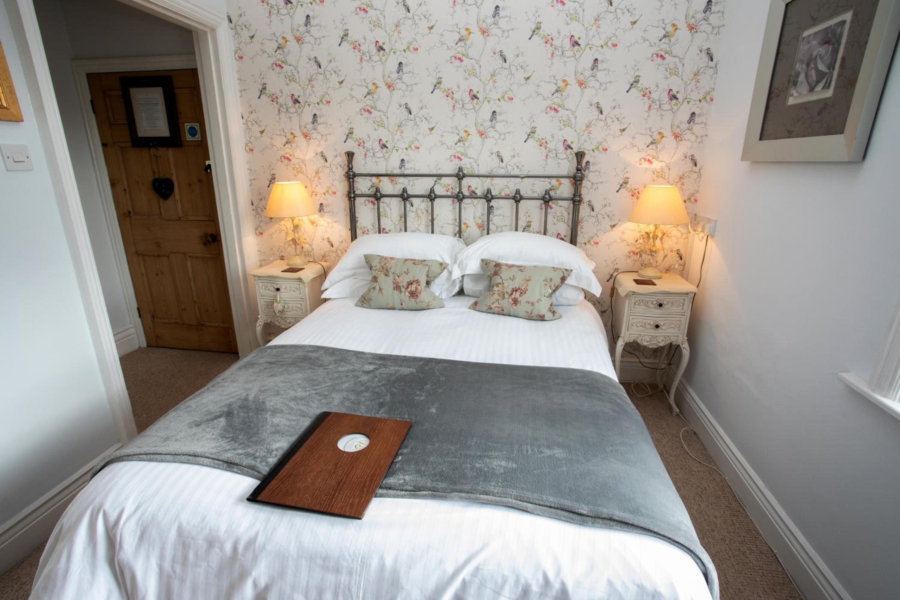 A bedroom with a bed, two nightstands, a lamp on each, a door, and a wall with floral wallpaper.