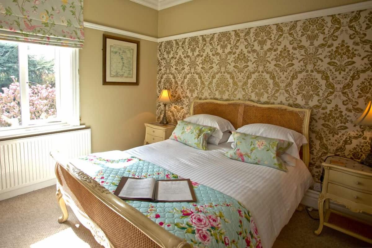Un dormitorio con una cama grande, ropa de cama floral, una mesita de noche con lámpara y una ventana con vistas a árboles.