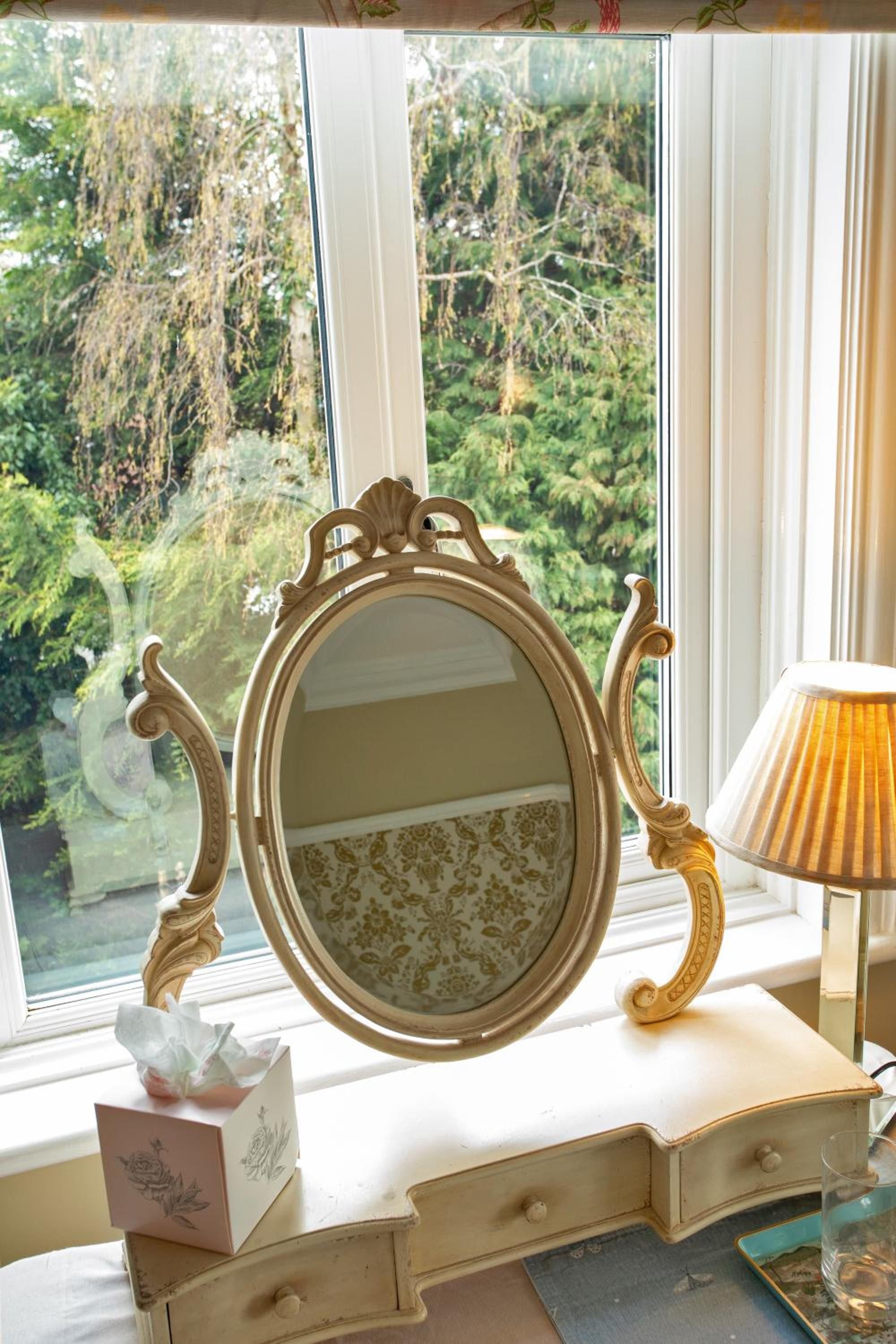 Un tocador de estilo vintage con espejo, un dispensador de pañuelos y una lámpara sobre un aparador junto a una ventana.