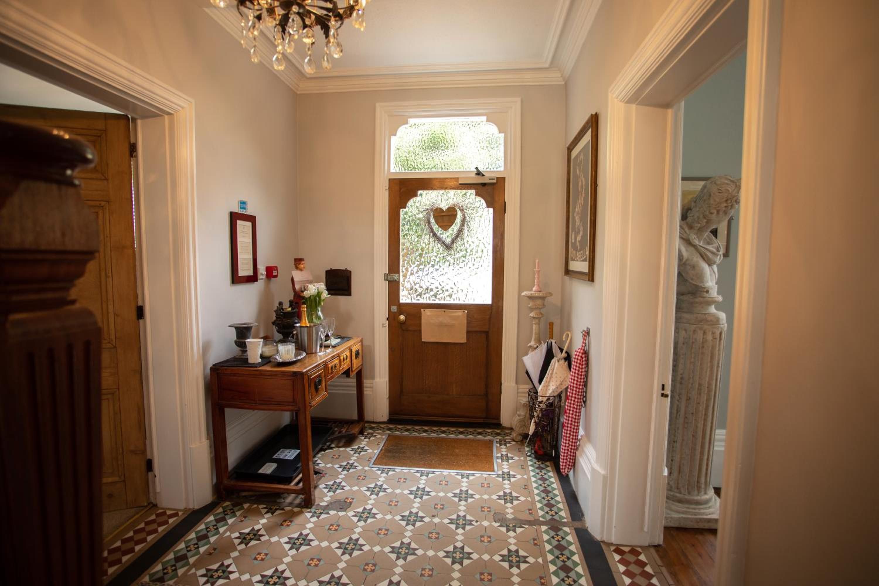 A hallway with a wooden door, a small table with a vase and glasses, a heart-shaped window, and a statue.