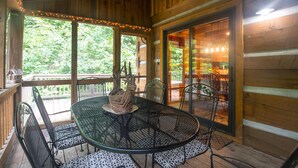 Eine Holzhütte mit einer Veranda, einem runden Tisch und Stühlen, großen Fenstern und Blick auf viel Grün.