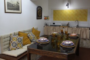Un comedor con una mesa de vidrio dispuesta para cuatro personas, un sofá con cojines estampados, una zona de cocina con estufa a gas y un salpicadero de azulejos decorativos.