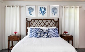 A bed with a white duvet, two pillows with blue and white patterns, and a wooden headboard. There are two nightstands with red flowers, and three framed wall art pieces above the bed. - Southern Breeze: Expansive Ocean Views! Amazing Sunrises! (St. John)