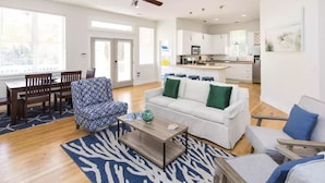 Una sala moderna con un sofá blanco, un sillón estampado y una mesa de centro. Hay un comedor con muebles de madera y un ventanal grande.
