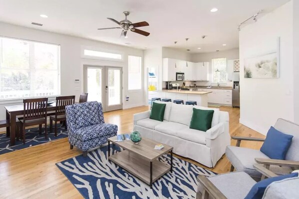 Una sala moderna con un sofá blanco, un sillón estampado y una mesa de centro. Hay un comedor con muebles de madera y un ventanal grande.