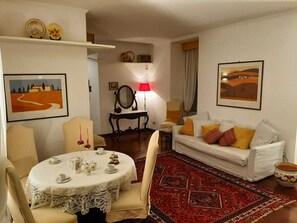 A living room with a round table set for tea, a sofa with colorful cushions, a red patterned rug, and two framed pictures on the walls.