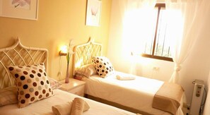 Two single beds with headboards and pillows feature in this cozy space, accompanied by a bedside table, lamp, and a vase with flowers.