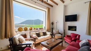 Una sala de estar con un ventanal grande, un sofá, un sillón rojo, una mesa de centro de madera y un televisor montado en la pared.